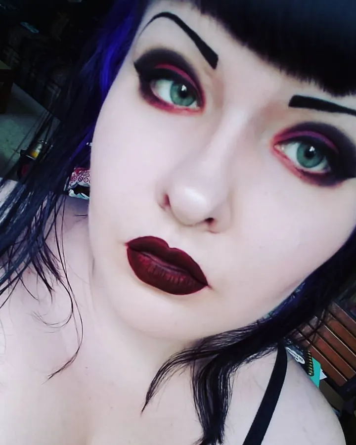 Femme trans aux cheveux bruns-noirs, en sous-vêtements noirs, posant sur un lit