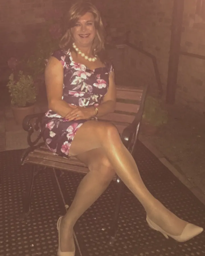 Femme trans aux cheveux blonds, en costume noir et jupe à carreaux, posant contre une barrière extérieure