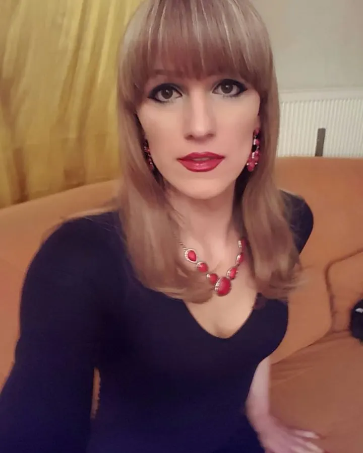 Jeune femme trans blonde, en robe jaune, posant sur un canapé blanc.