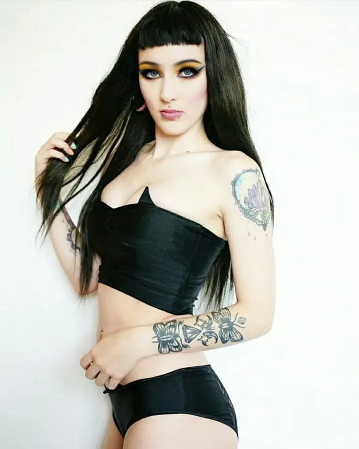 Femme trans aux cheveux bruns-noirs, en haut noir, regardant l'objectif, torse nu, photo en couleur, ambiance intime.