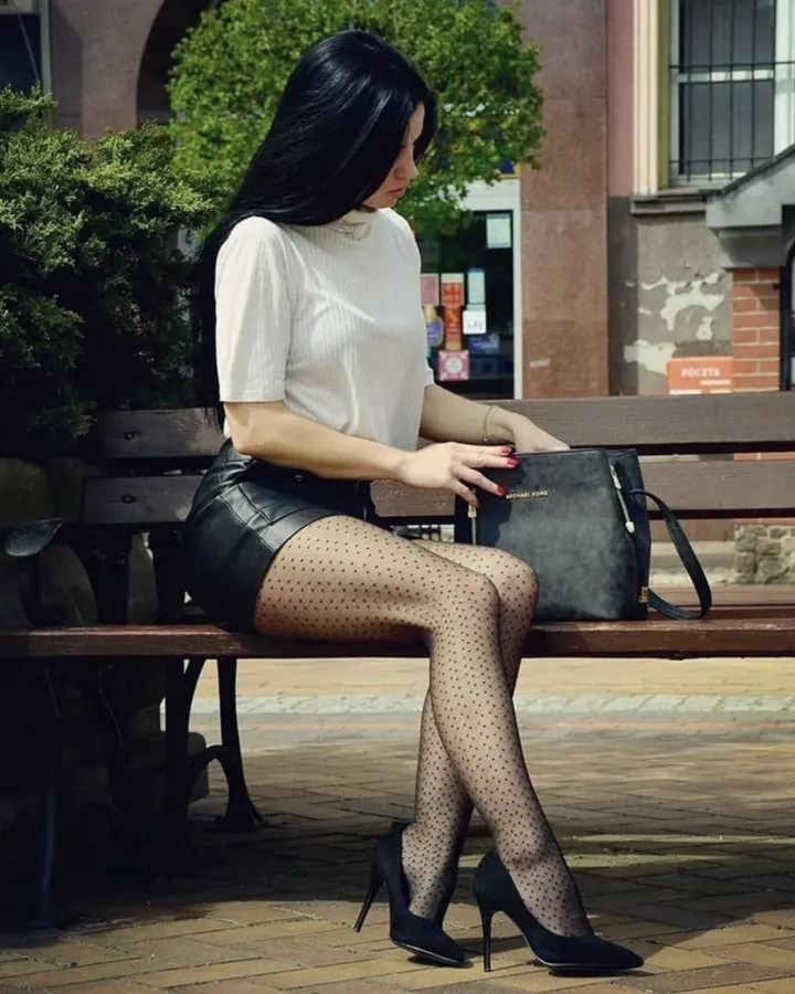 Femme trans aux cheveux bruns-noirs, en haut blanc et jupe en cuir noir, assise sur un banc en bois en ville, portant des bas à pois et des talons noirs.