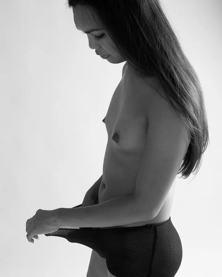 Femme trans aux cheveux bruns-noirs, en haut noir, regardant l'objectif, torse nu, photo en couleur, ambiance intime.