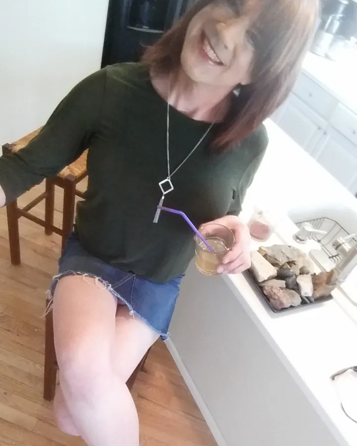 Femme trans aux cheveux blonds, en haut vert foncé et jupe en jean courte, assise sur un tabouret, tenant un verre avec paille violette, souriant largement.