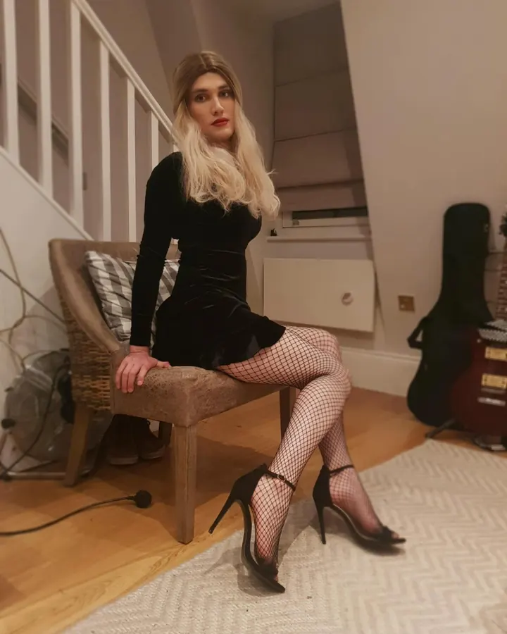 Femme trans aux cheveux blonds, en robe noire velours, collants résille, chaussures à talons, assise sur une chaise dans un intérieur moderne.