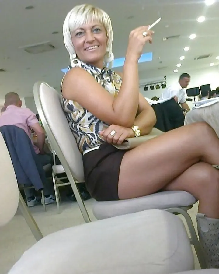 Femme trans européenne aux cheveux platine, assise sur une chaise blanche, tenant une cigarette, souriant, dans un restaurant ou une salle de conférence.