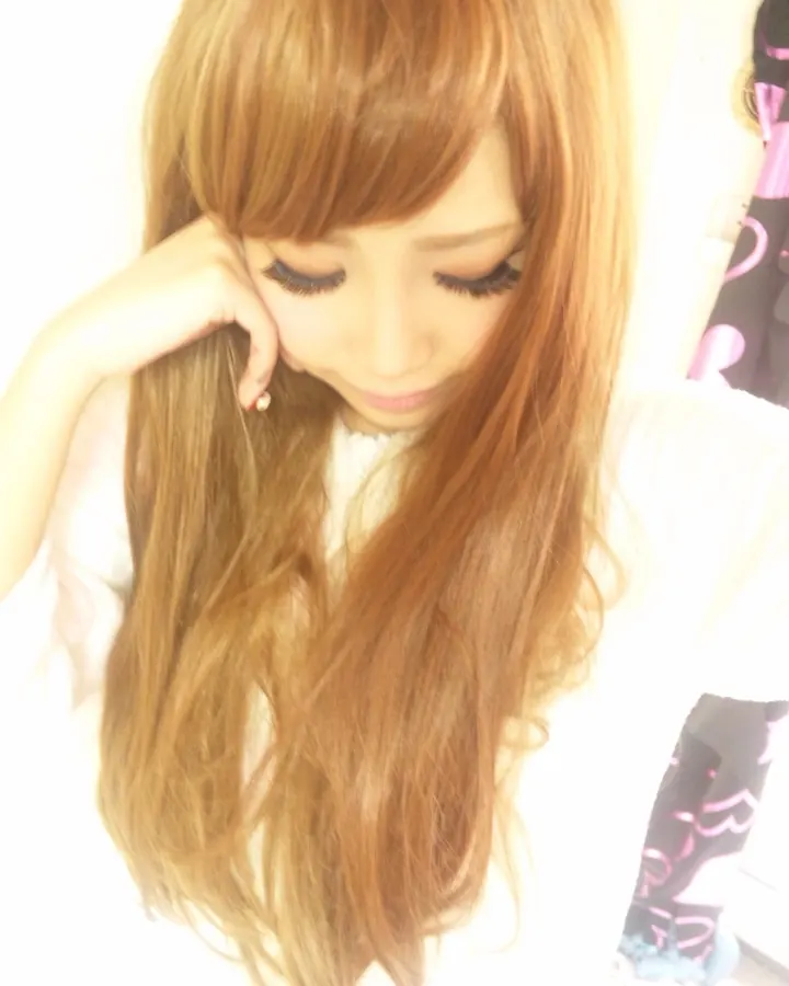Femme trans asiatique aux cheveux blonds, portant un haut noir, se tenant debout dans une chambre, tenant un téléphone pour prendre un selfie.