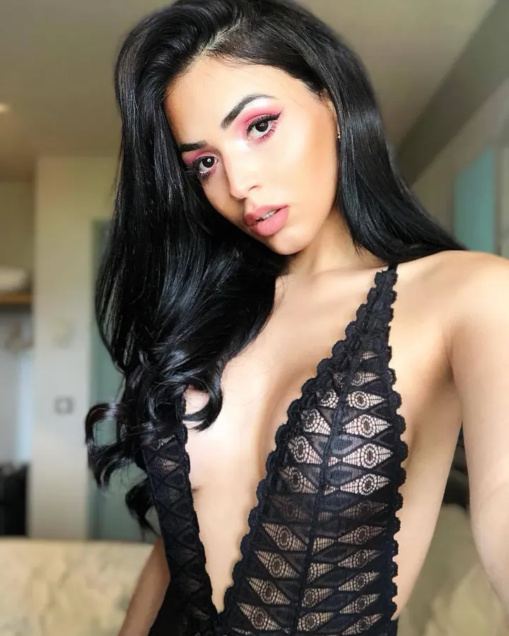 Femme trans aux cheveux bruns, torse nu, posant sur un canapé beige, avec une main sur son entrejambe.