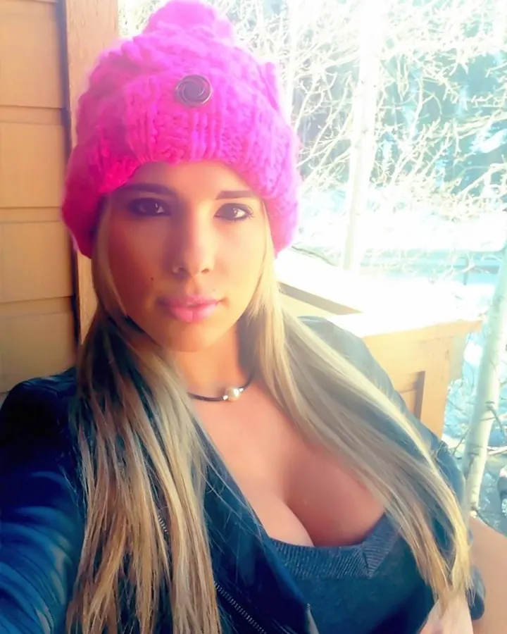 Femme trans aux cheveux blonds, portant un bonnet rose, posant en extérieur, avec une veste en cuir.
