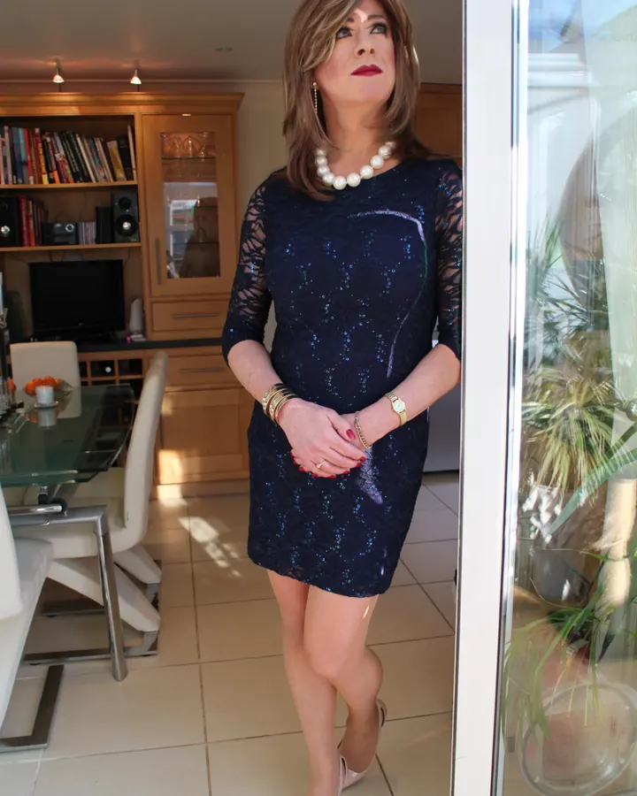 Femme trans aux cheveux blonds, portant une robe bleue scintillante, une collier de perles, posant dans un salon moderne.