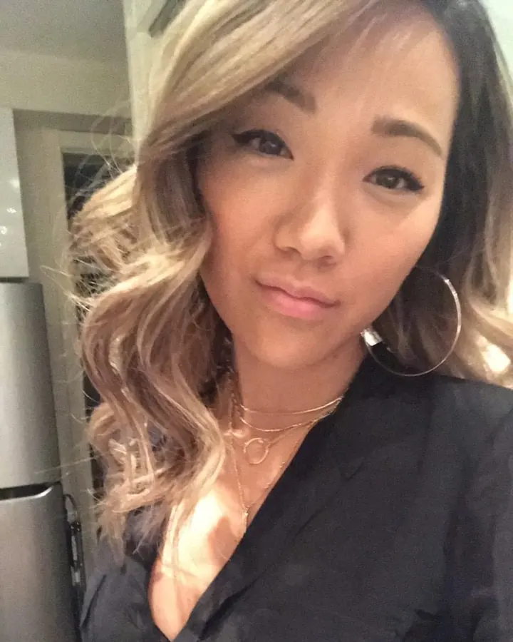 Jeune femme trans asiatique aux cheveux blonds, prenant un selfie dans une cuisine, regardant l'objectif avec un sourire discret