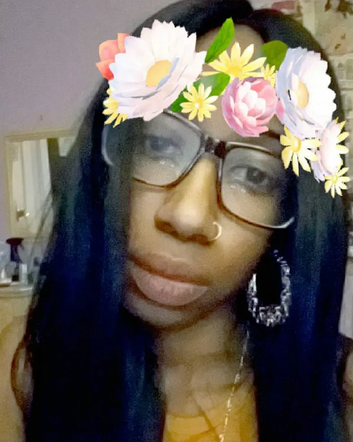 Femme trans aux cheveux noirs portant des lunettes et un filtre floral sur la tête, souriant légèrement.
