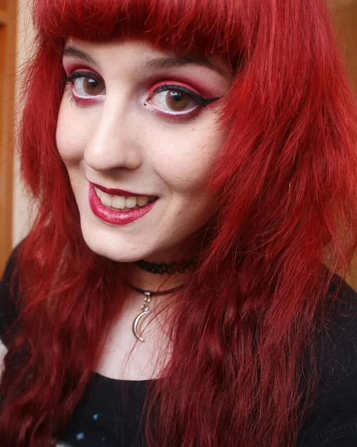 Femme trans aux cheveux roux portant un haut noir, souriant avec un collier lunaire.