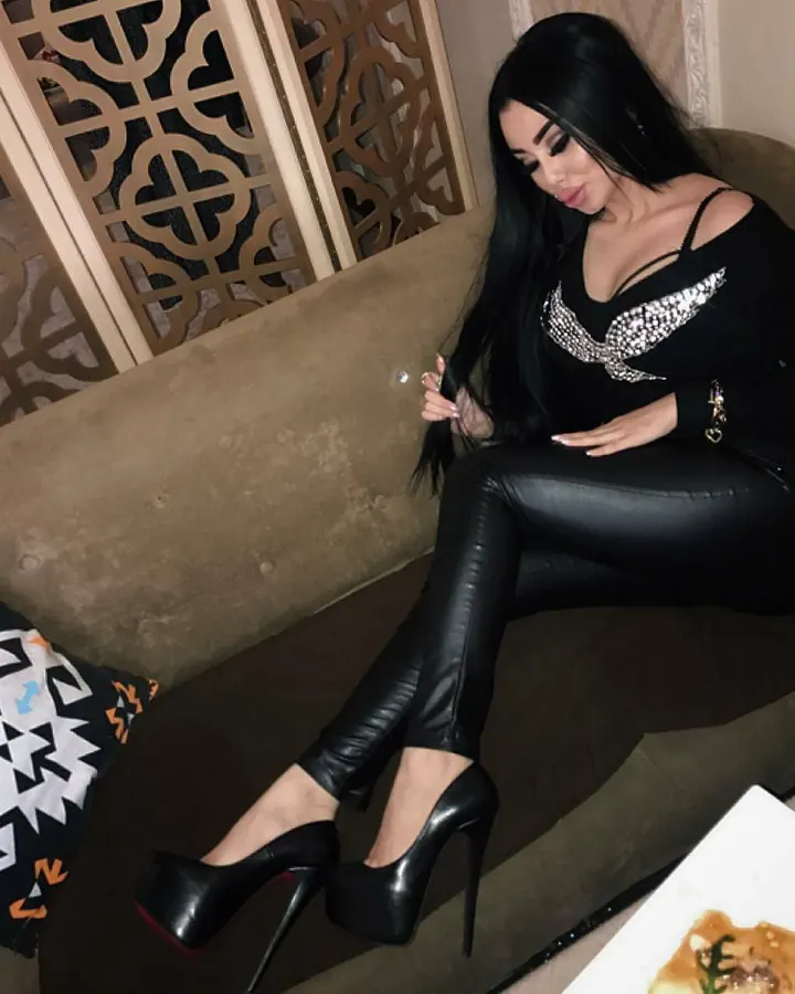 Femme trans aux cheveux noirs, portant un haut noir et un pantalon en cuir noir, assise sur un canapé beige avec des talons noirs à plateforme.