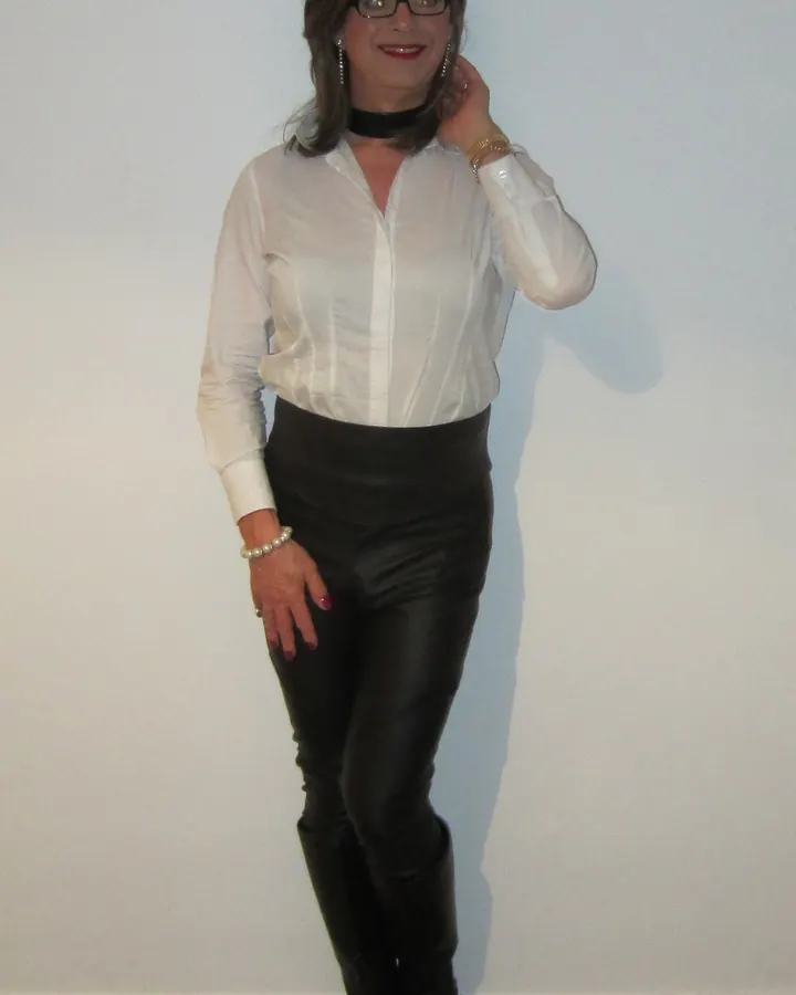 Femme trans aux cheveux bruns, portant une robe noire transparente, des collants noirs, des chaussures noires et des bijoux de perles, assise sur un canapé marron dans un restaurant.