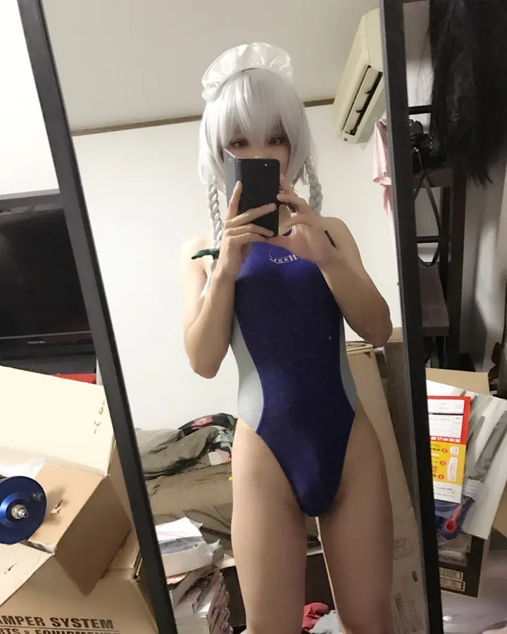 Personne aux cheveux blancs en maillot de bain bleu et blanc, prenant un selfie devant un miroir dans une pièce encombrée.