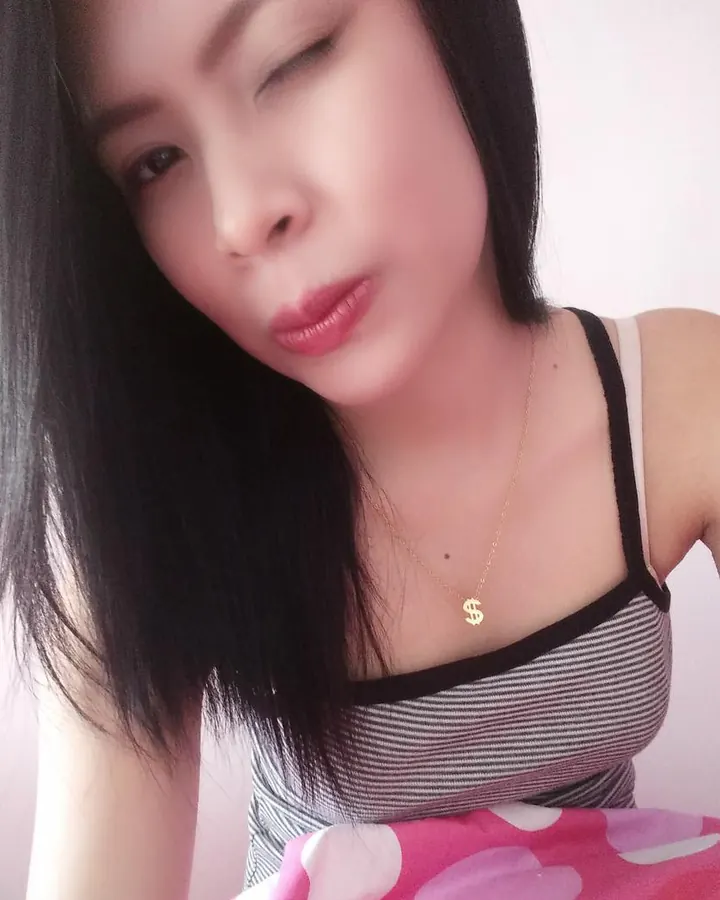 Femme trans aux cheveux noirs, portant un haut noir, avec un collier en cuir et des boucles d'oreilles en chaîne, prenant un selfie.