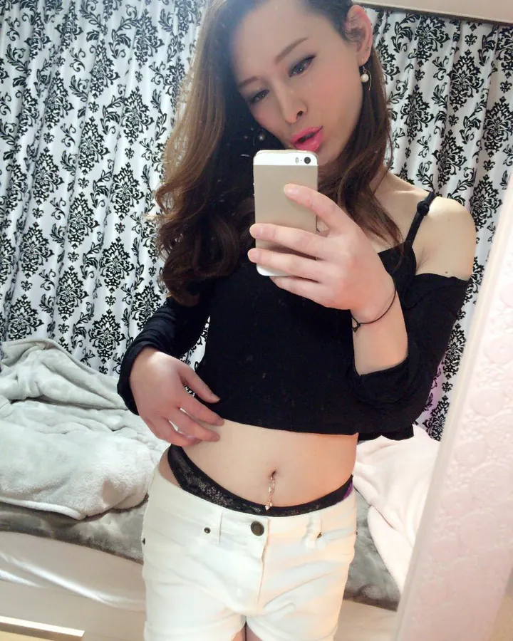 Femme trans aux cheveux bruns, portant un haut noir et un short blanc, prenant un selfie devant un miroir avec un rideau à motifs noirs et blancs en arrière-plan.