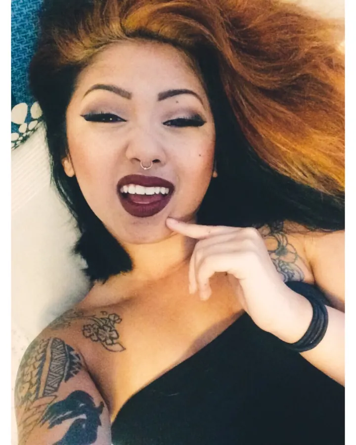 Femme trans aux cheveux blonds, en lingerie blanche, posant avec des tatouages visibles sur les bras.
