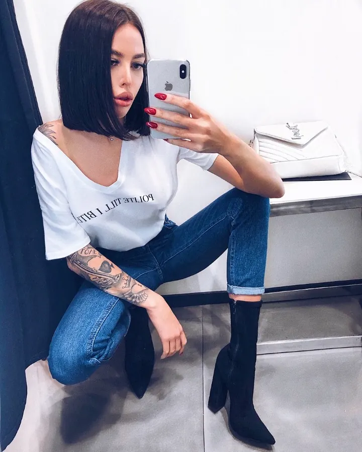 Femme trans aux cheveux bruns-noirs, en lingerie blanche, posant avec des tatouages visibles sur les bras.